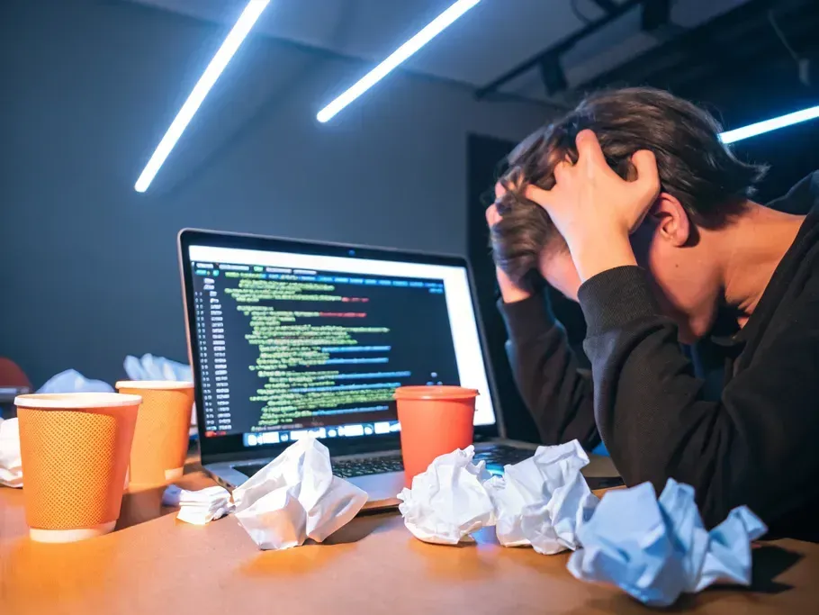 Person frustrated at a computer screen filled with code, surrounded by crumpled paper and empty coffee cups.