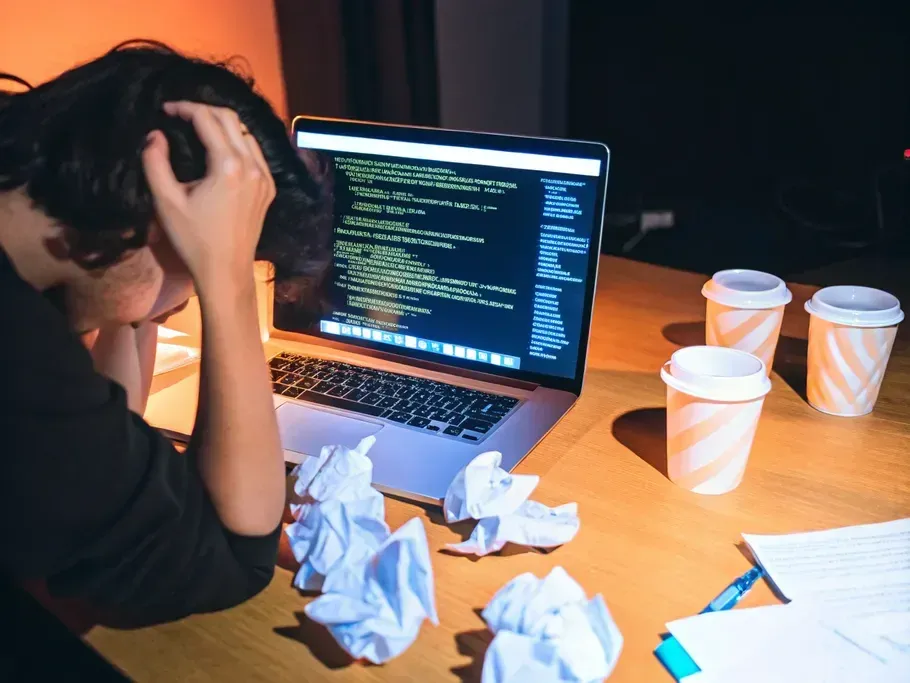 A frustrated person at a computer displaying incomprehensible code, surrounded by crumpled paper and empty coffee cups.