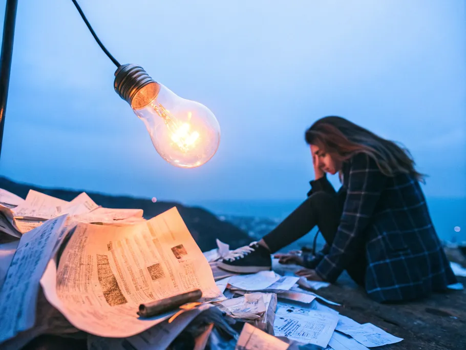 A woman sitting near a pile of papers under a glowing light bulb at dusk, symbolizing innovation in grief technology