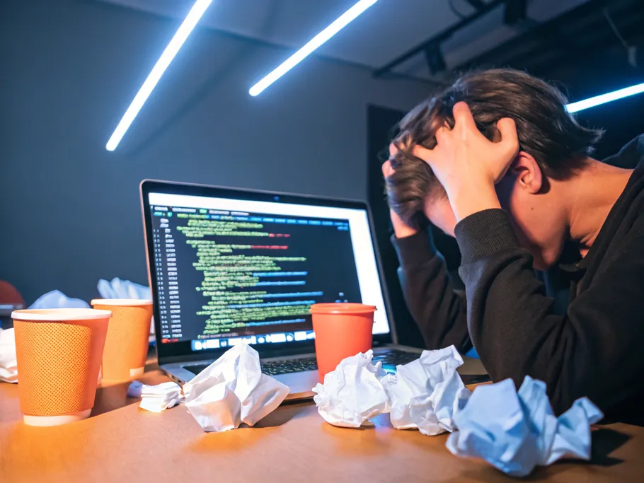 Person frustrated at a computer screen filled with code, surrounded by crumpled paper and empty coffee cups.