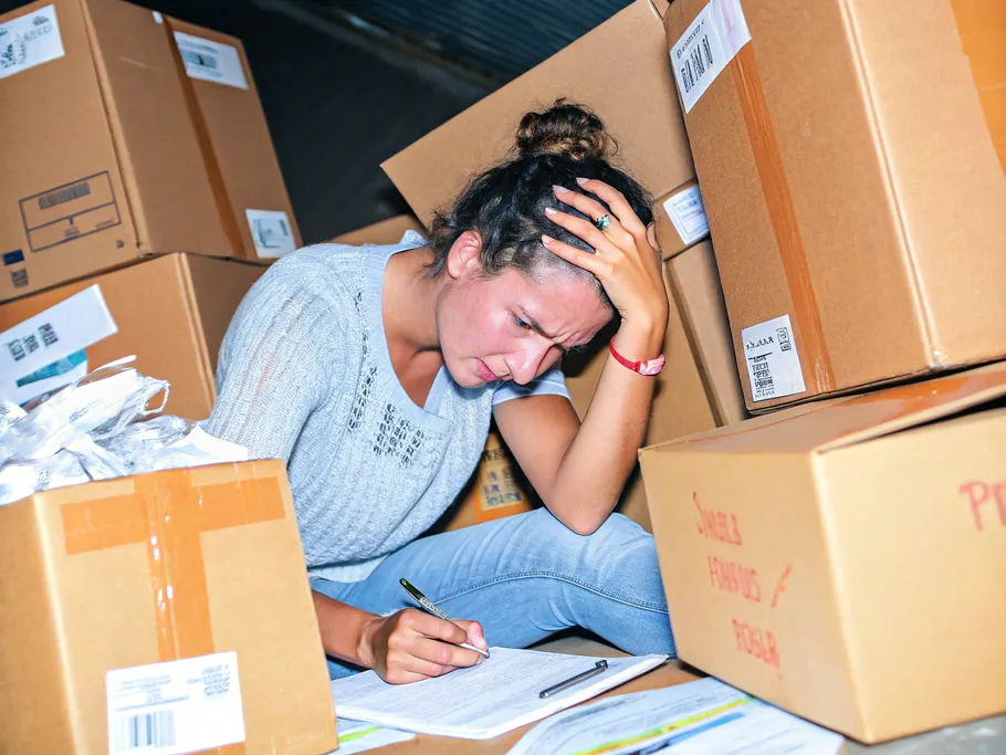 A young artisan overwhelmed by a mountain of cardboard boxes, packing tape, and shipping labels, looking stressed and exhausted.