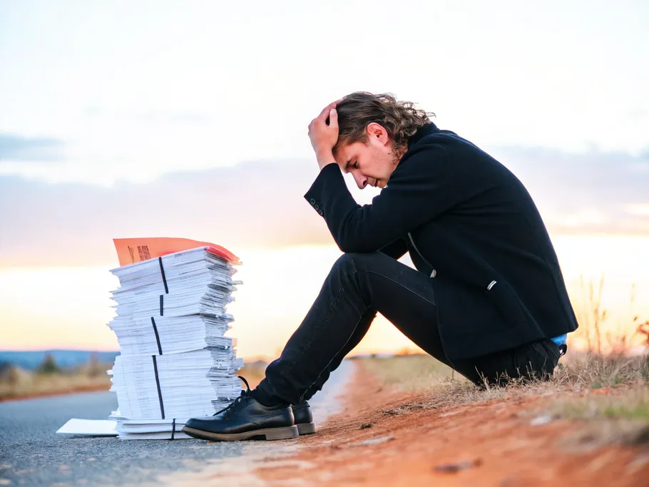 A young startup employee seated beside a stack of financial documents, expressing a mixture of burden and hope.